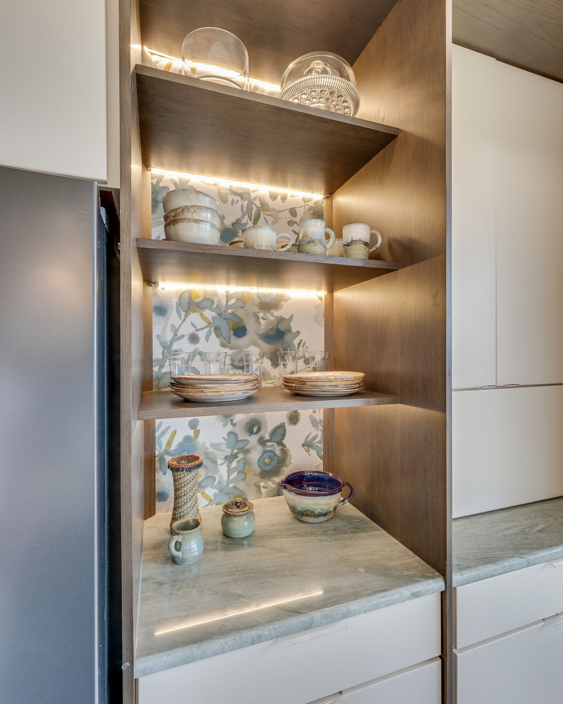 Built-in kitchen shelves with lit china, floral plates, and teacups above white cabinets.
