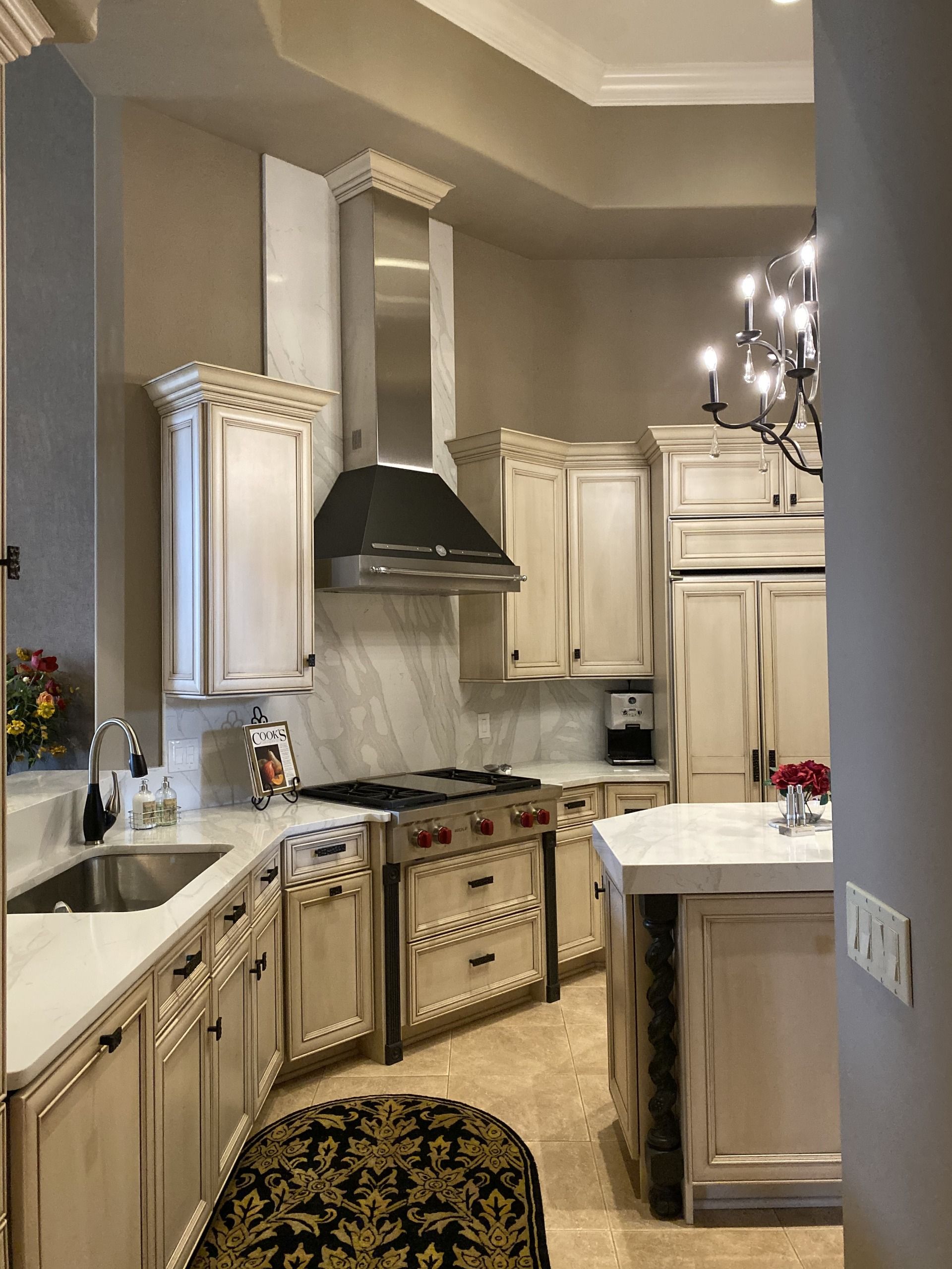 Elegant cream-colored kitchen with marble counters, black range hood, and chandelier lighting