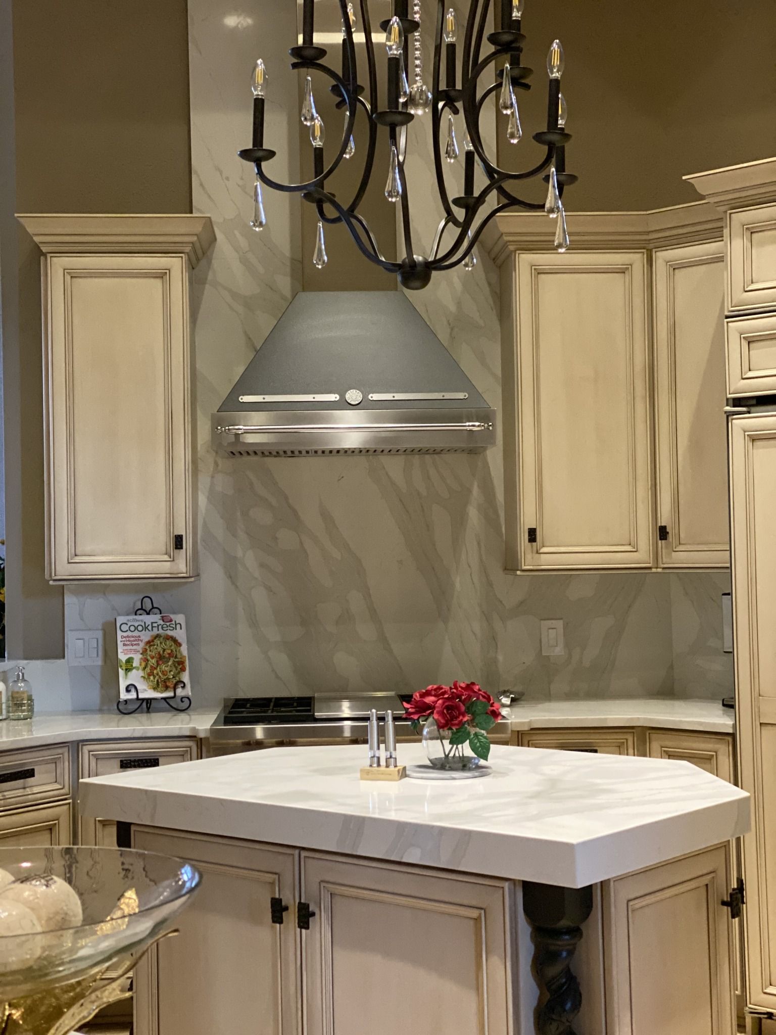 Elegant kitchen with cream cabinets, marble island, stainless hood, and black chandelier above a flower centerpiece.