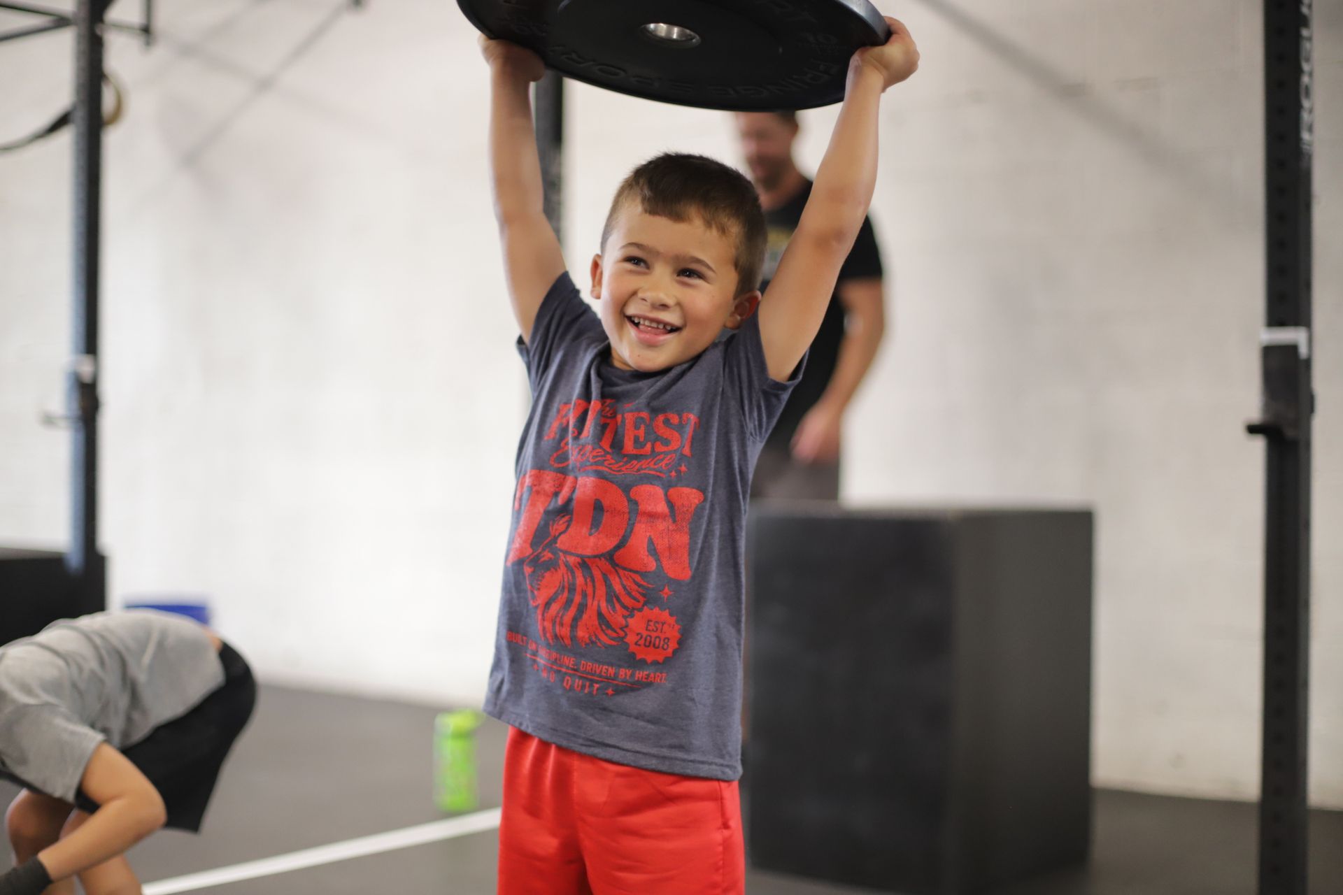 Smiling child in a black “PUPPY DAWGS” shirt posing in a gym with exercise bikes at CrossFit Exile