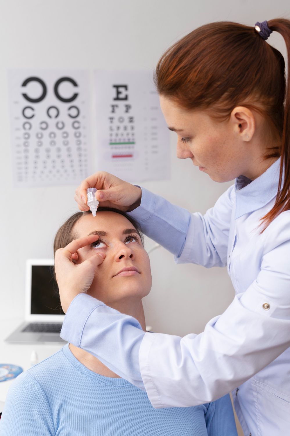 Optometrist administering eye drops to a patient in an exam room, eye chart in the background.