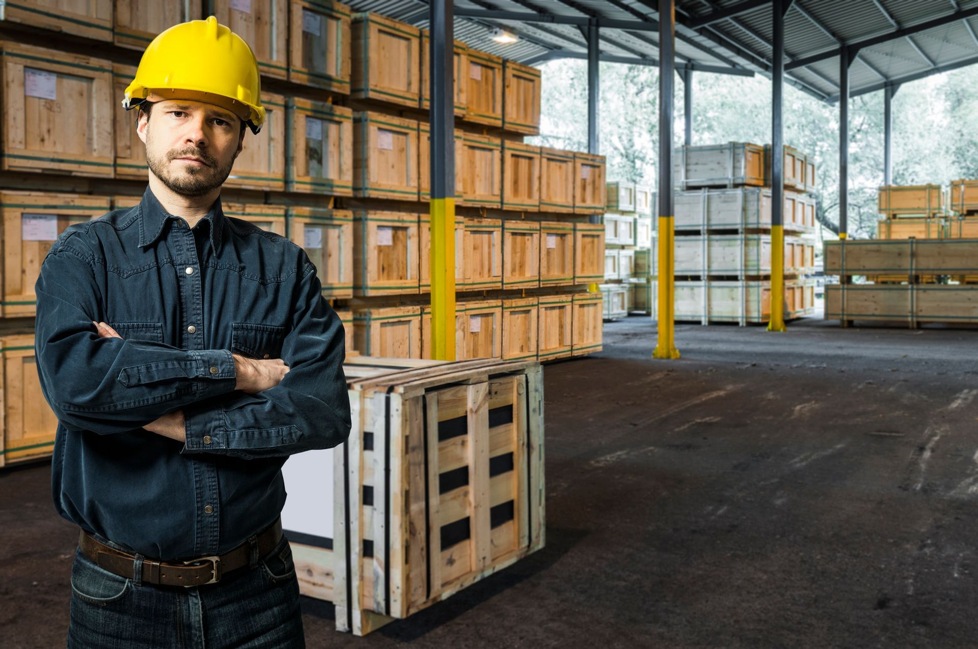 A man wearing a hard hat is standing in a warehouse with his arms crossed.