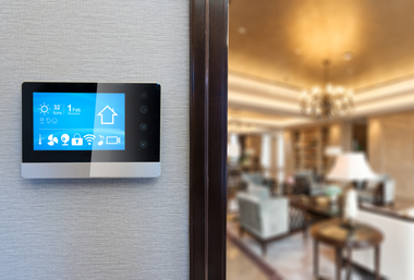 Wall-mounted touchscreen thermostat beside a blurred living room with warm lighting