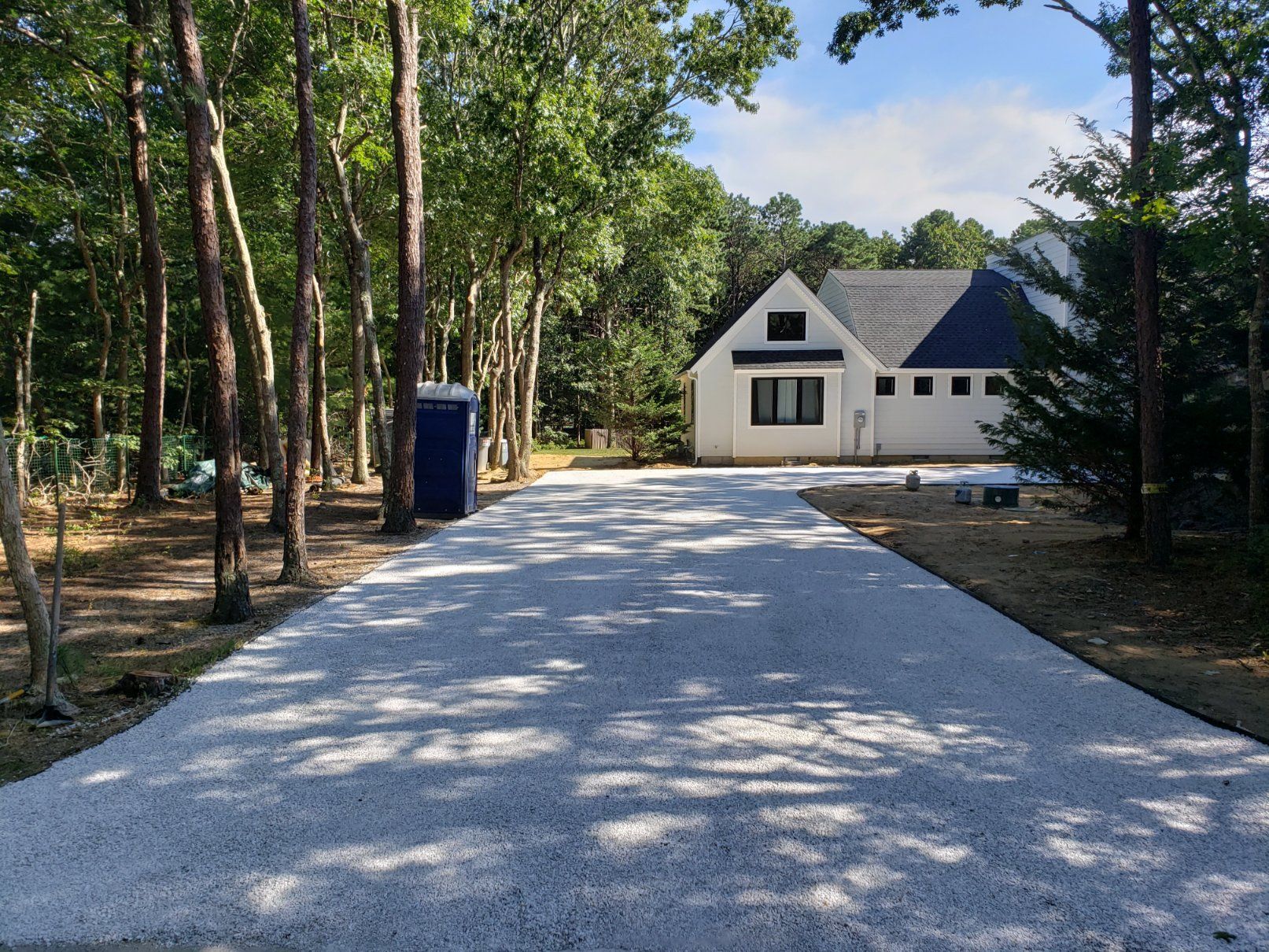 A gravel driveway leads through a wooded area toward a white house with a dark gabled roof and a portable toilet nearby.