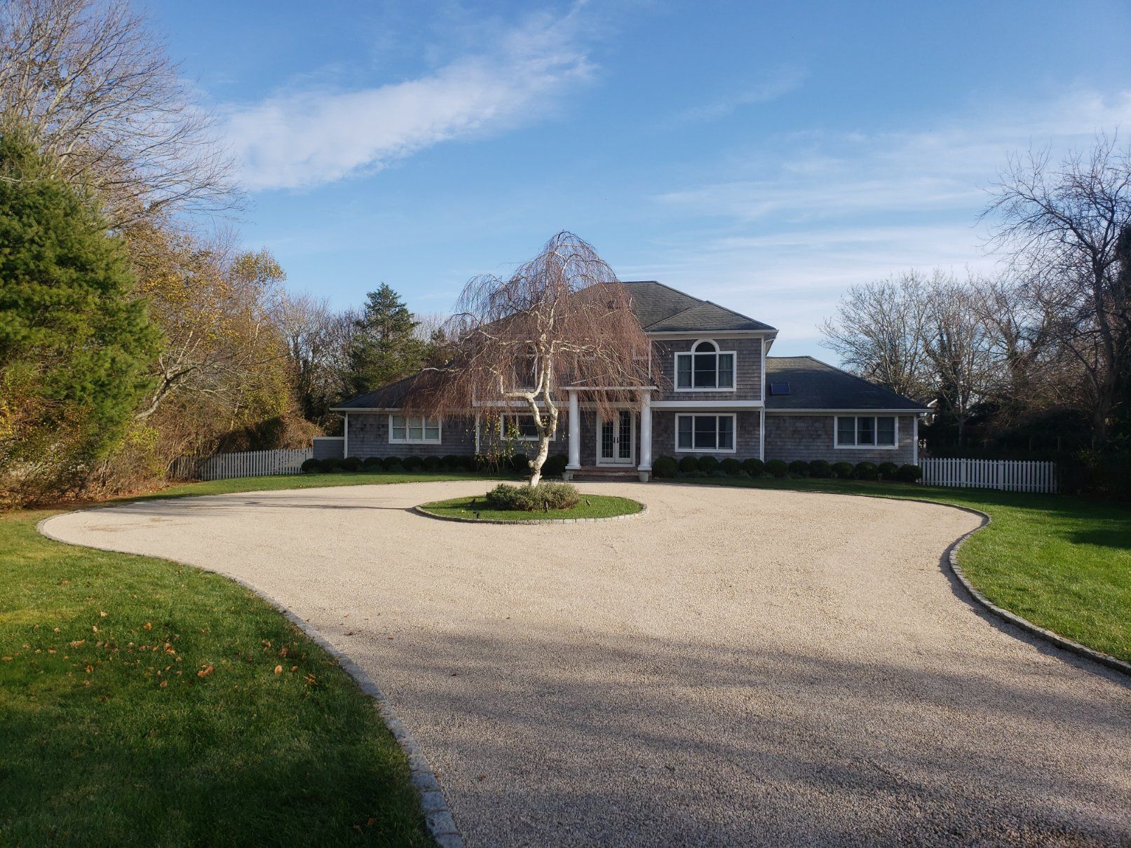 A two-story grey shingled house behind a large gravel driveway with a small central tree, surrounded by grass and trees.