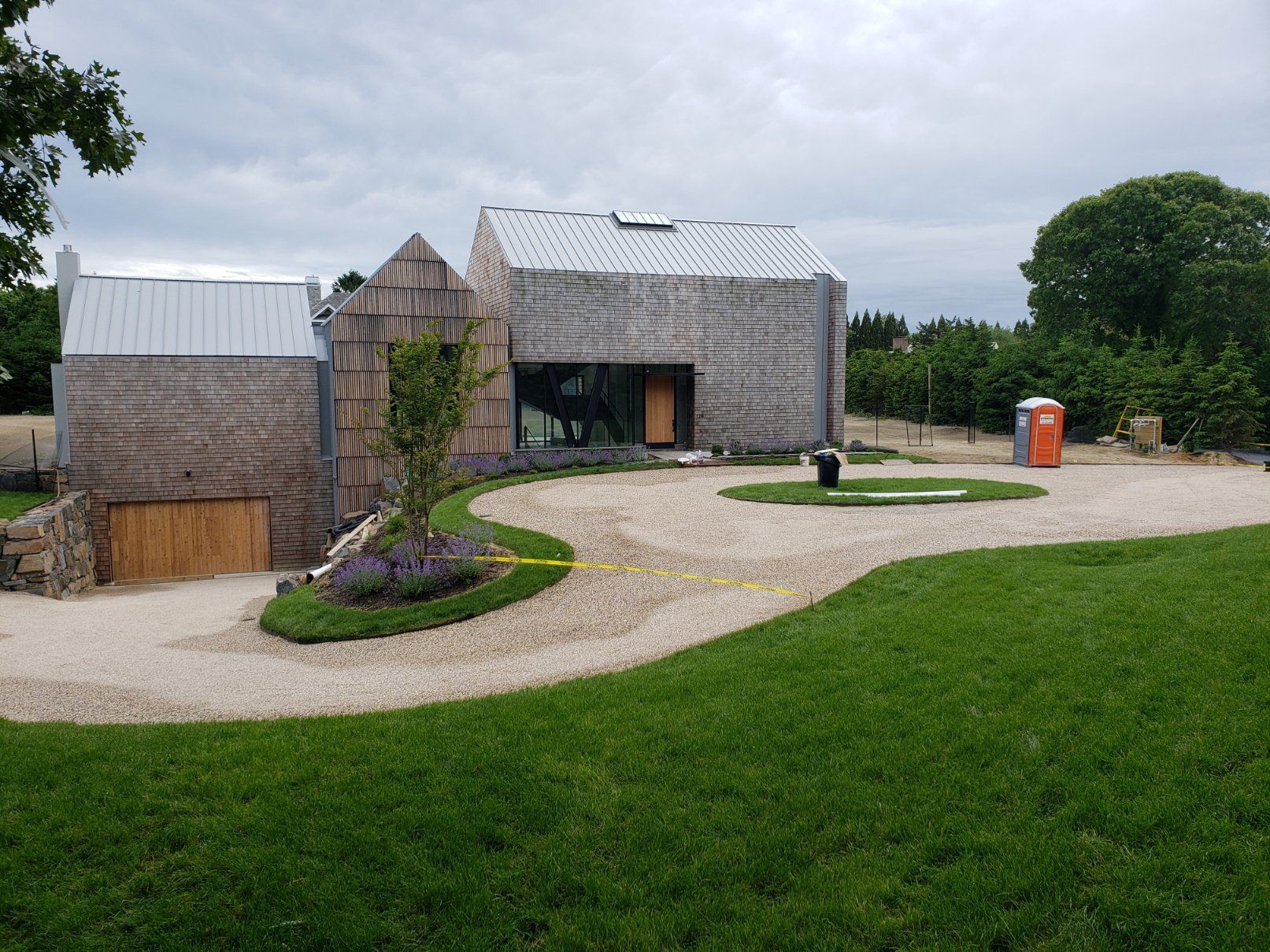 A modern house with shingled walls and a metal roof sits behind a curved gravel driveway on a lawn.