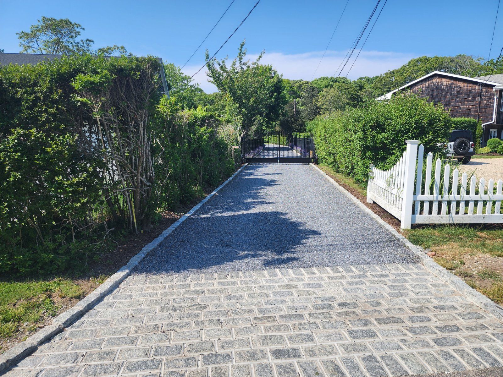 A driveway with a cobblestone apron transitioning to a gravel path, lined by a green hedge and a white picket fence.
