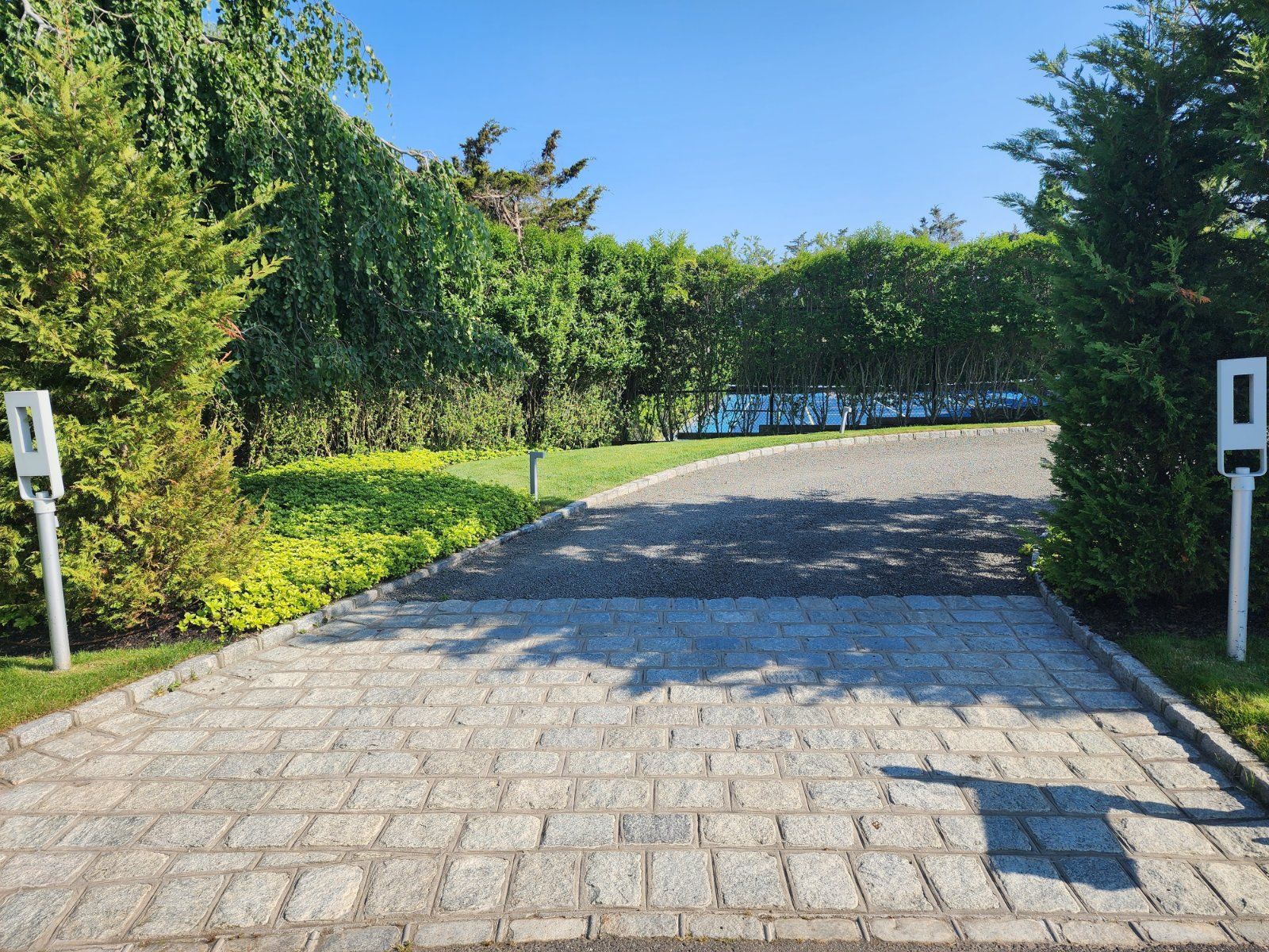 A stone driveway framed by two white posts leads into a lush, green landscaped area under a clear blue sky.