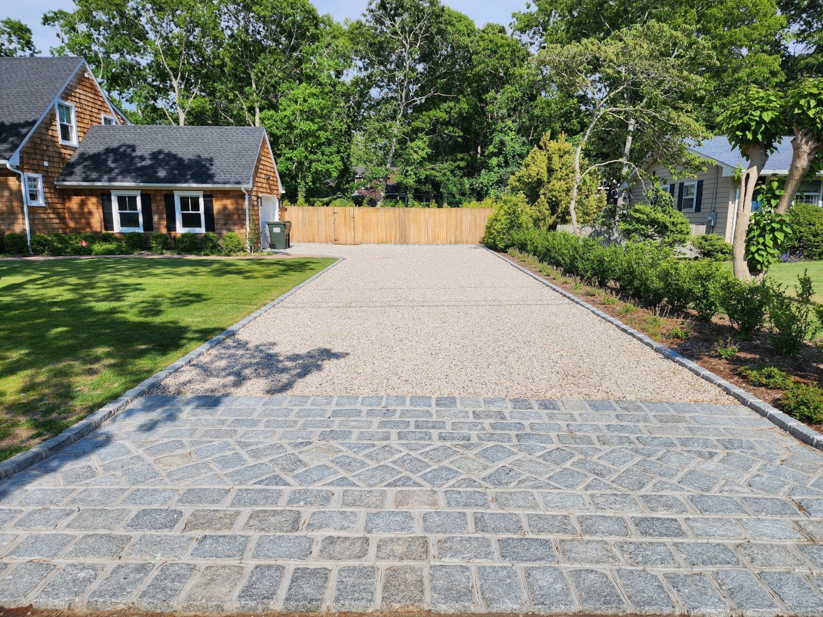 A gravel driveway with a patterned cobblestone apron leading to a house, flanked by a lawn and a hedge on a sunny day.
