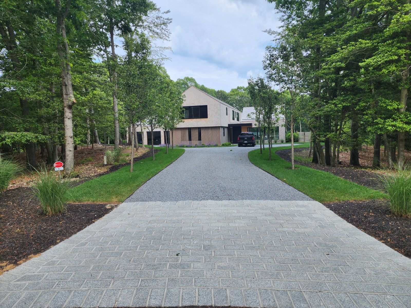 A paved driveway leads to a modern home nestled in a wooded area with trees lining both sides.