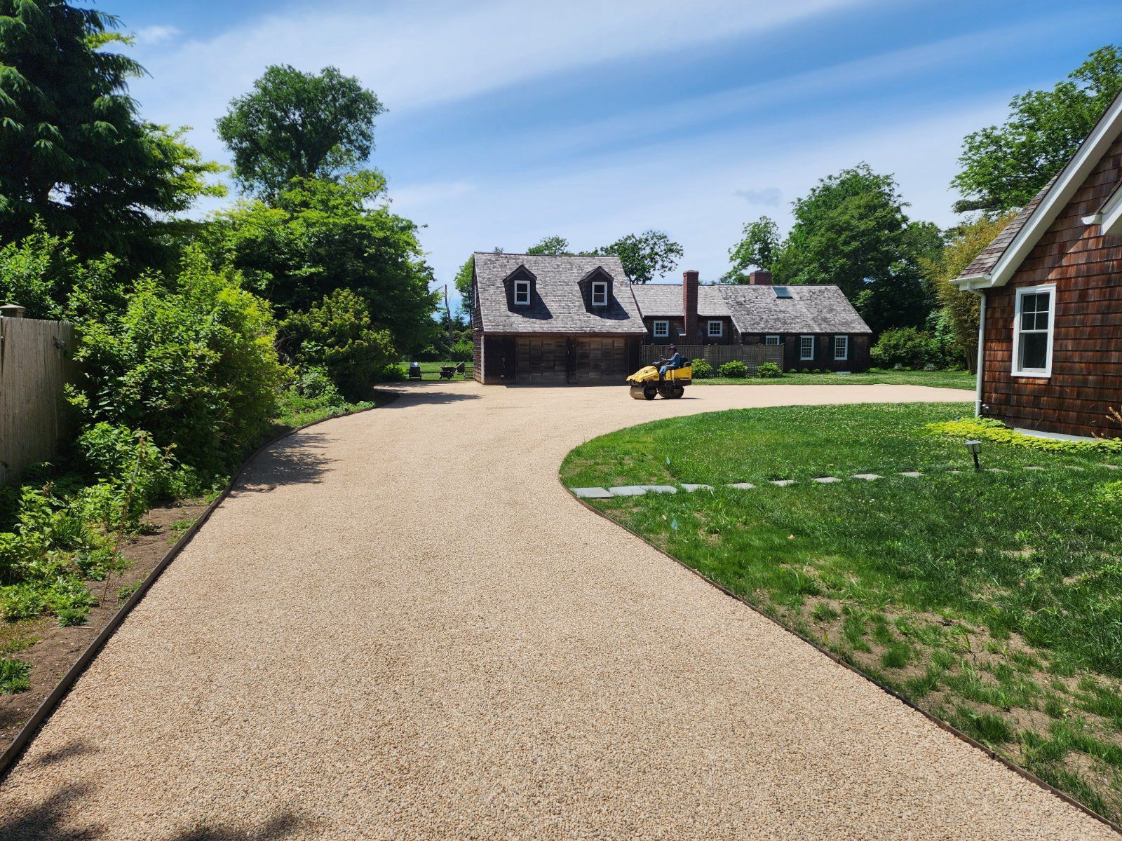 A tan gravel driveway leads to a gray-roofed house on a sunny day with green lawns and trees.