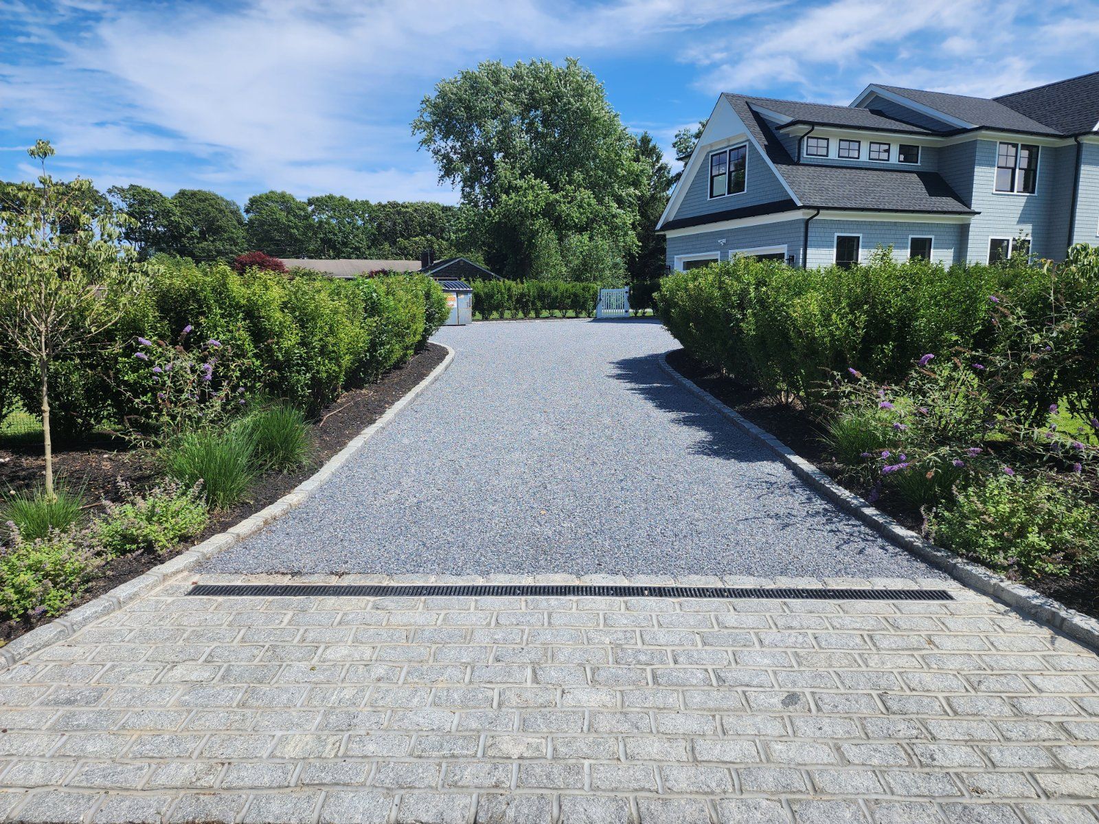 A driveway transition from gray stone pavers to a gravel surface, lined by manicured green hedges leading to a house.
