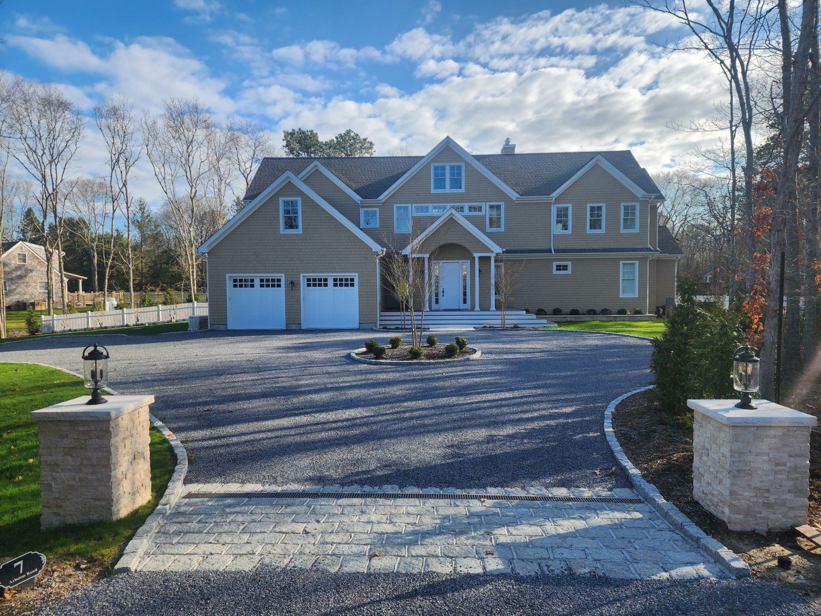 Tan two-story house with a white garage and door, viewed from a large gravel driveway with stone pillar entrance markers.