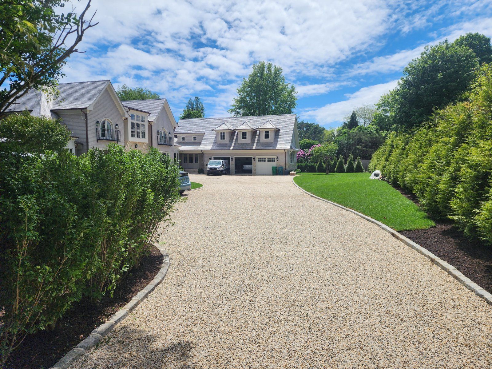 A gravel driveway leads to a light-colored multi-story house with a garage, flanked by green hedges and a grassy lawn.
