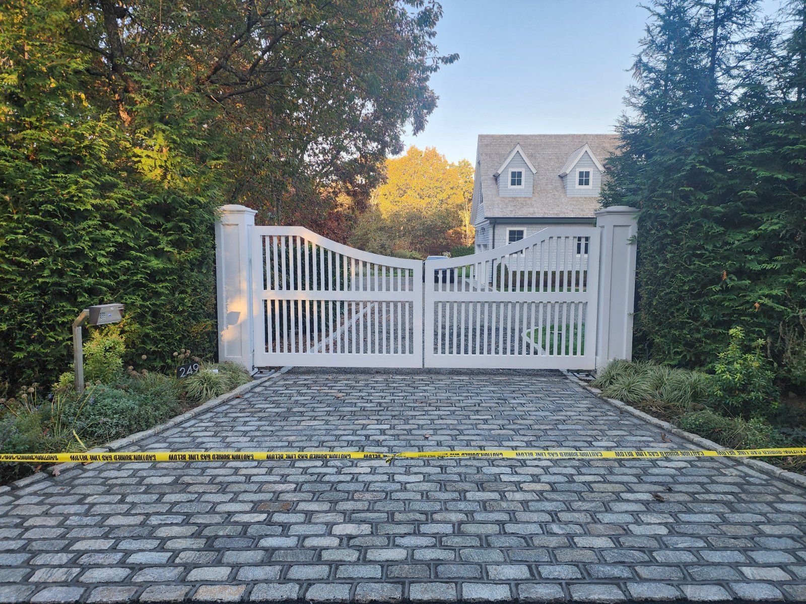 A closed white wooden gate with a cobblestone driveway in front, marked by yellow caution tape.