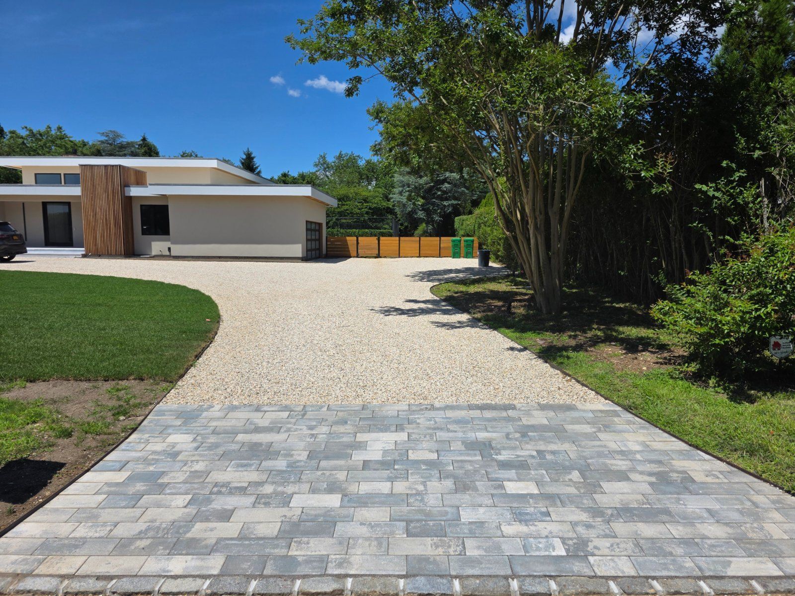 A driveway with gray stone pavers transitioning to light gravel, leading to a modern house on a sunny day.