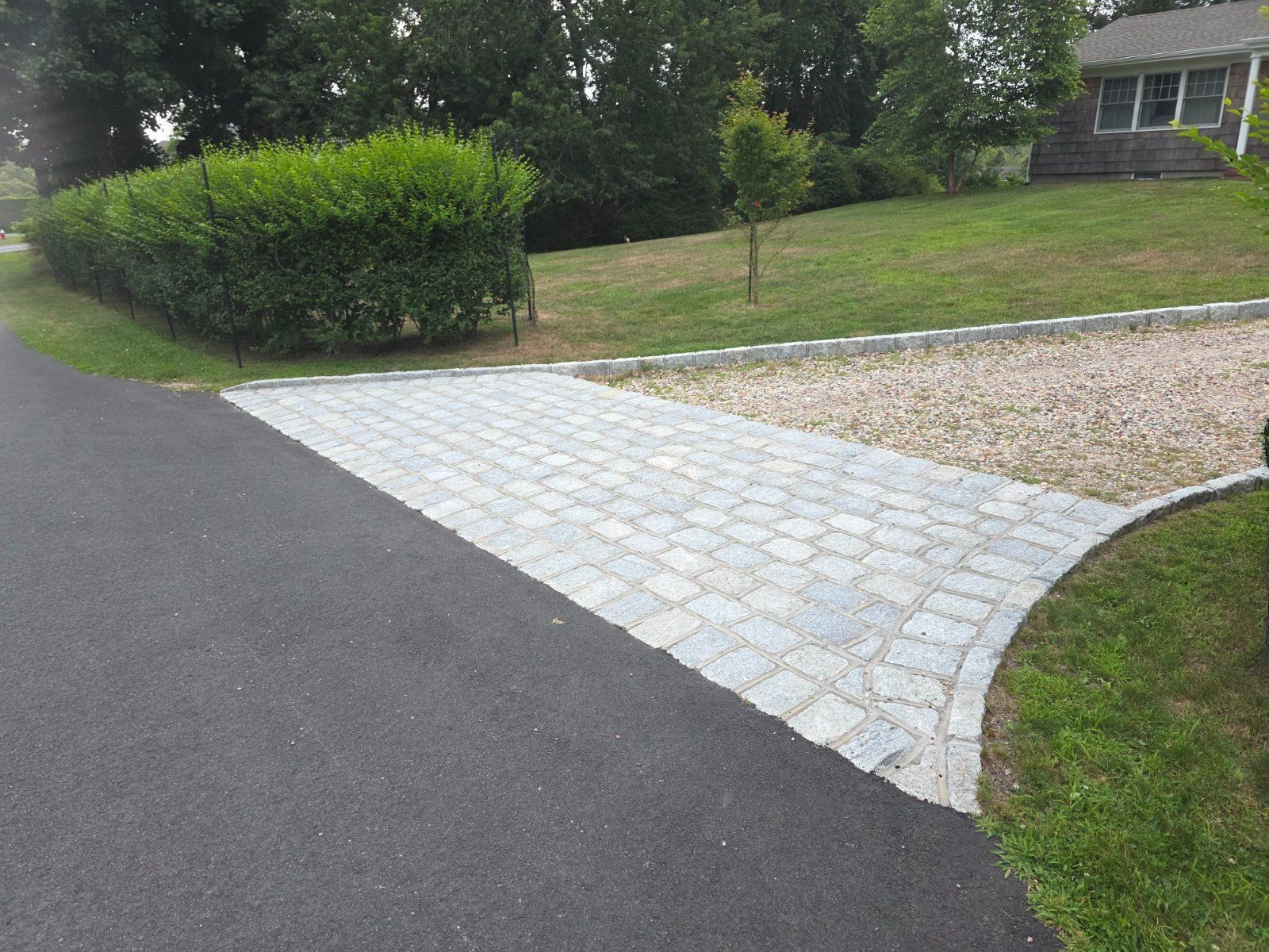 A stone paver driveway apron connects an asphalt road to a gravel driveway leading toward a house.