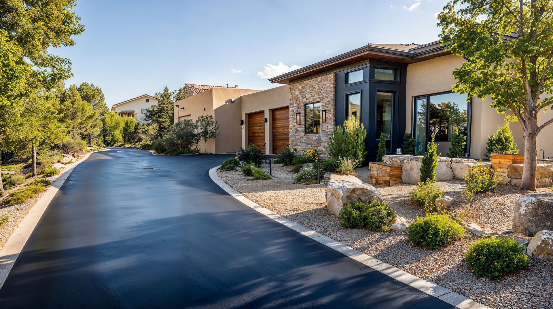 A paved driveway curves toward a modern stucco home with stone accents, wood garage doors, and landscaped desert terrain.