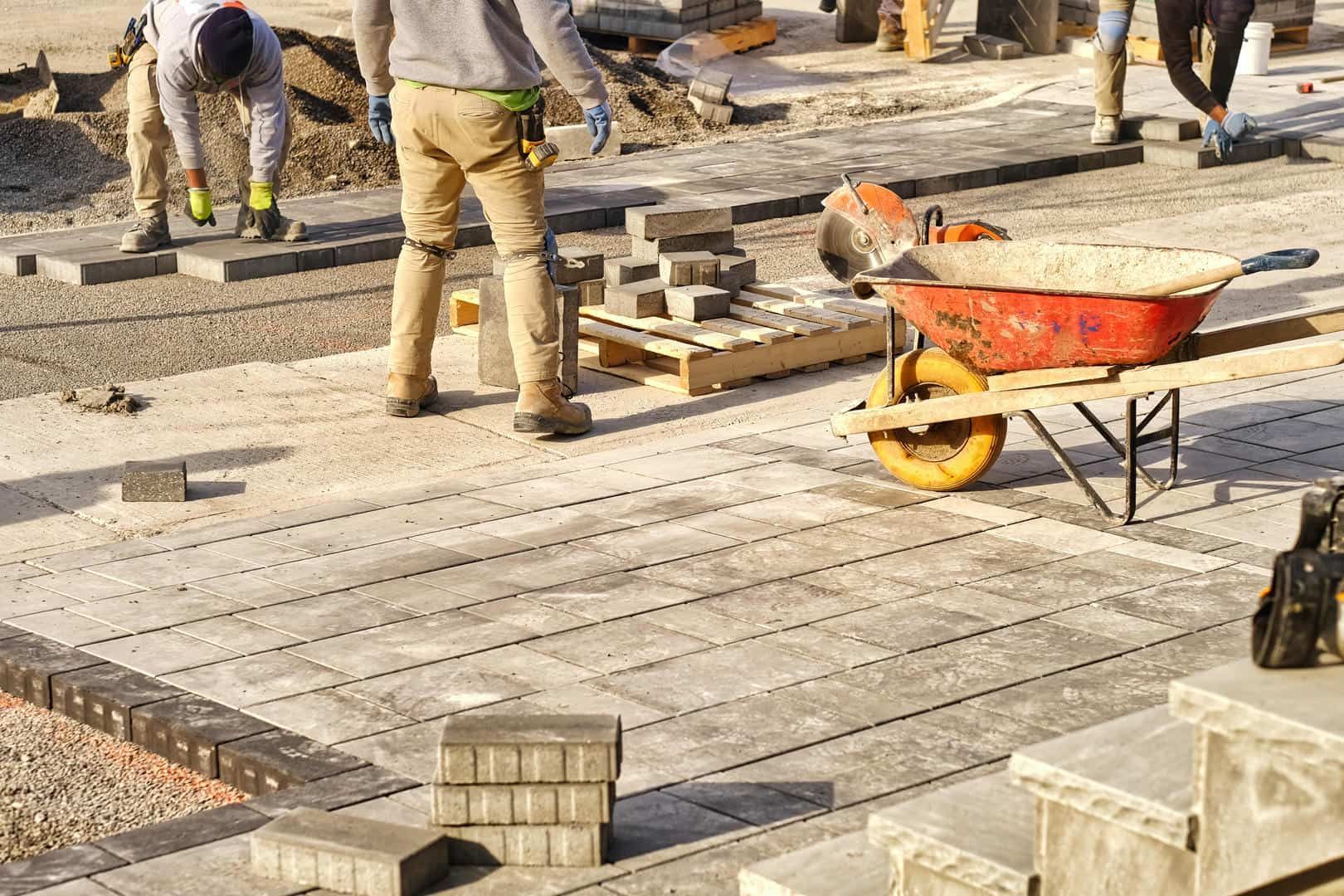Two workers in work clothes laying concrete pavers on a sandy ground near a wheelbarrow.
