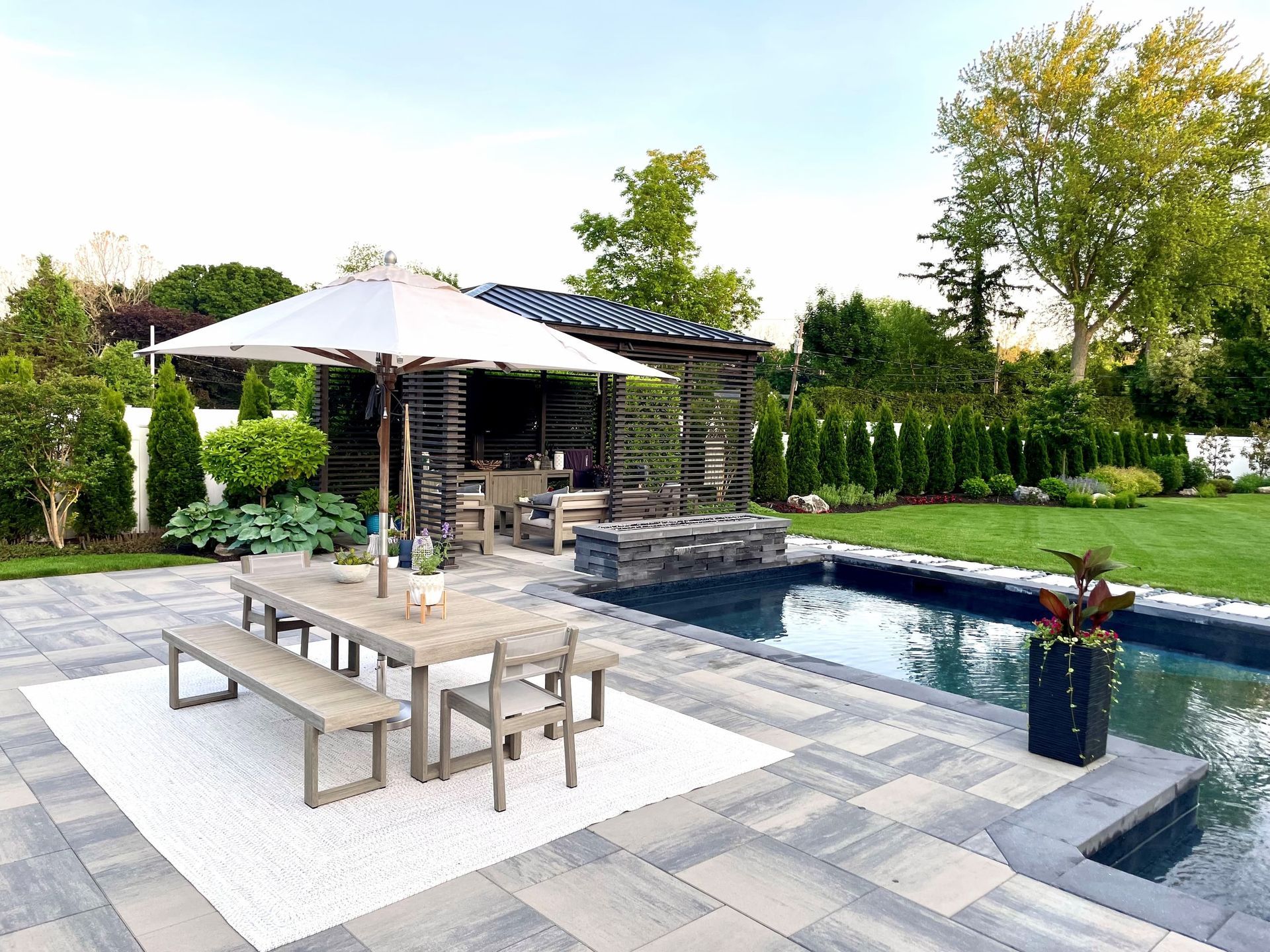 A wooden dining set with an umbrella on a white patio rug next to a swimming pool and a covered outdoor lounge area.