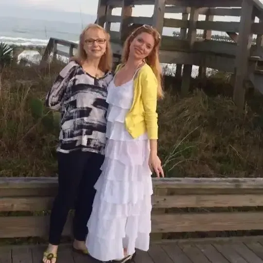 Two women stand on a boardwalk by the ocean in a white tiered dress, yellow sweater
