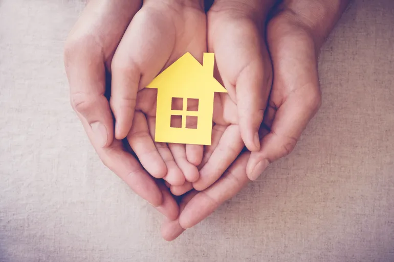 Hands cupping a small yellow paper house, representing care or protection.