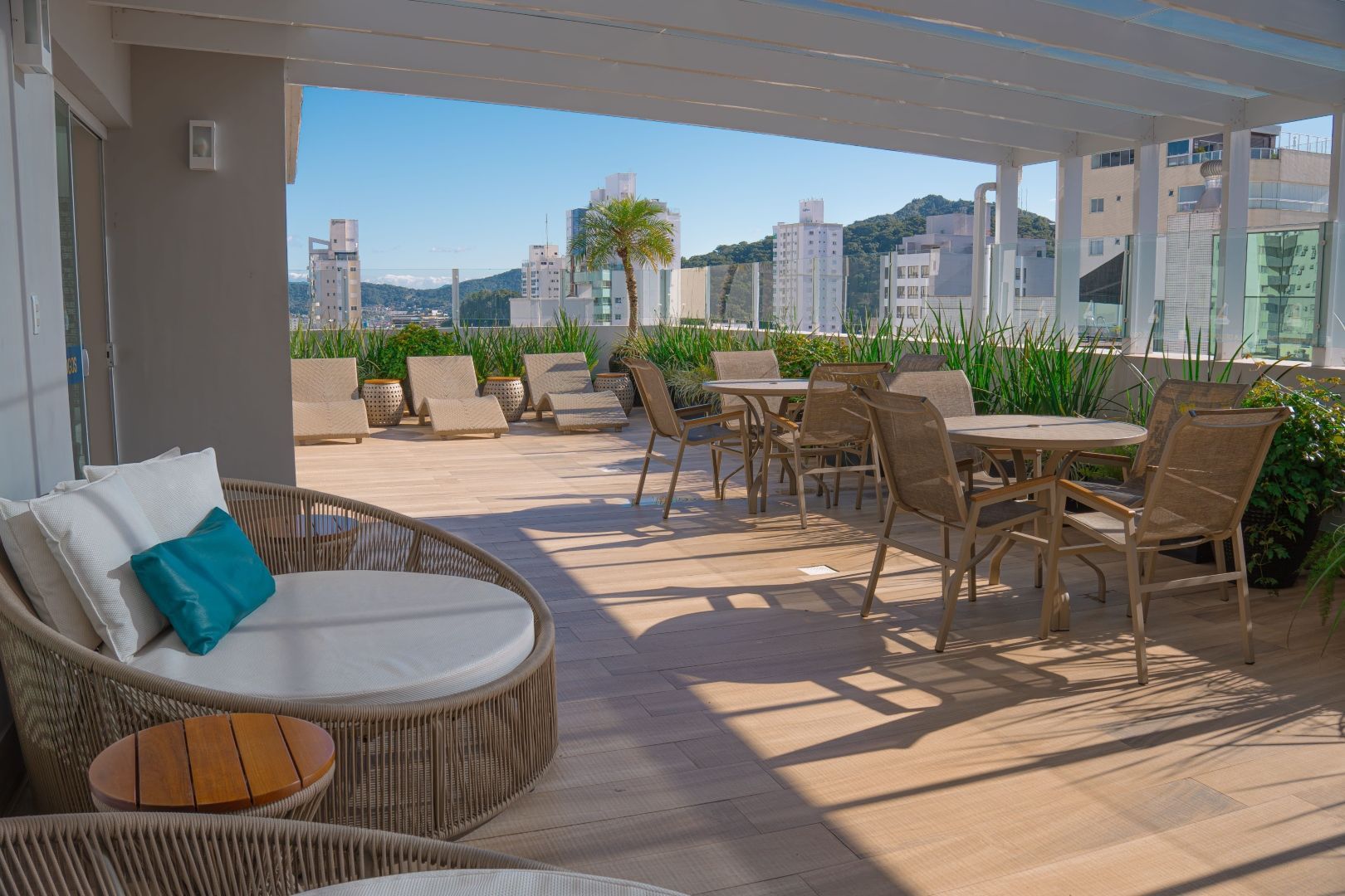 Terraço com espreguiçadeiras, mesas e vista para a cidade sob um céu ensolarado.