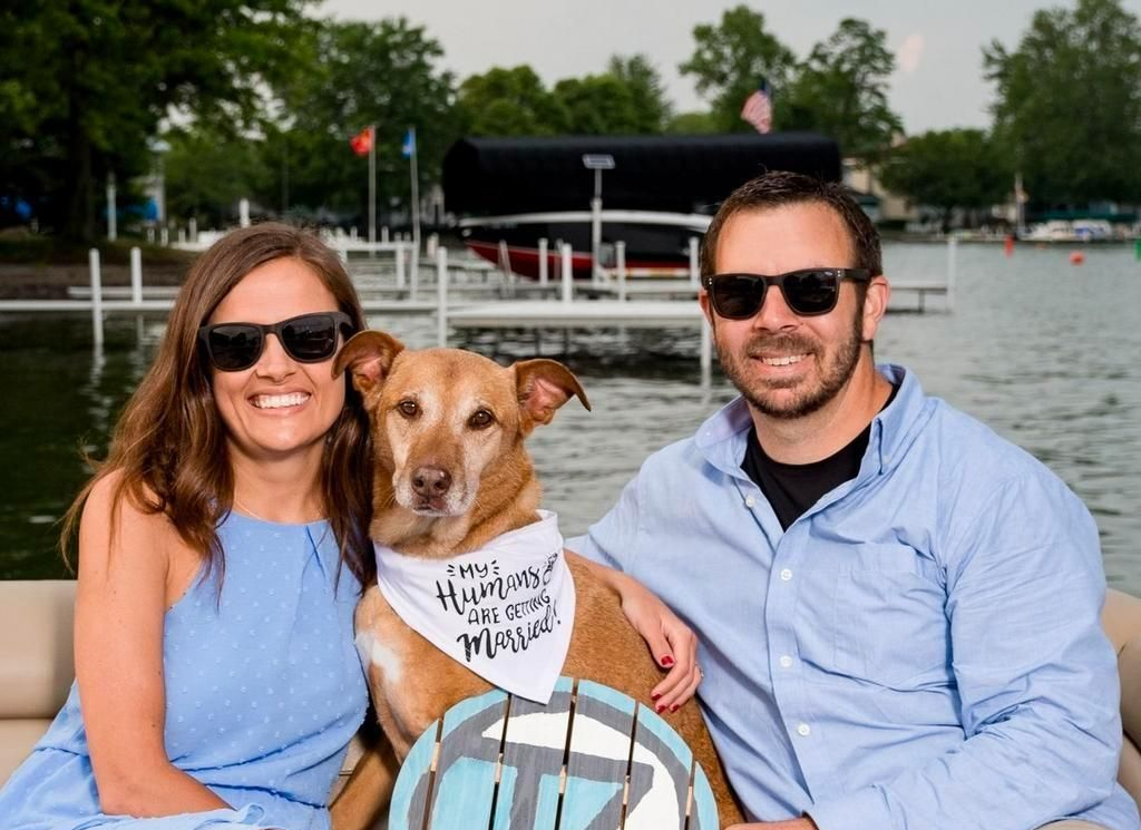 Smiling couple with a dog in a bandana sitting by the water on a boat.
