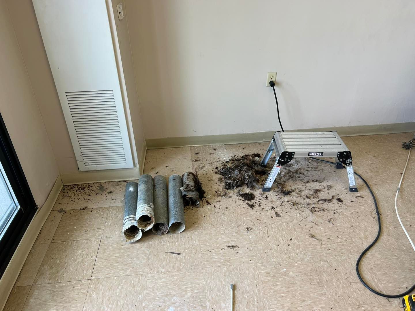 Room with debris, rolled metal pipes, and a small work table near a wall outlet and cord.
