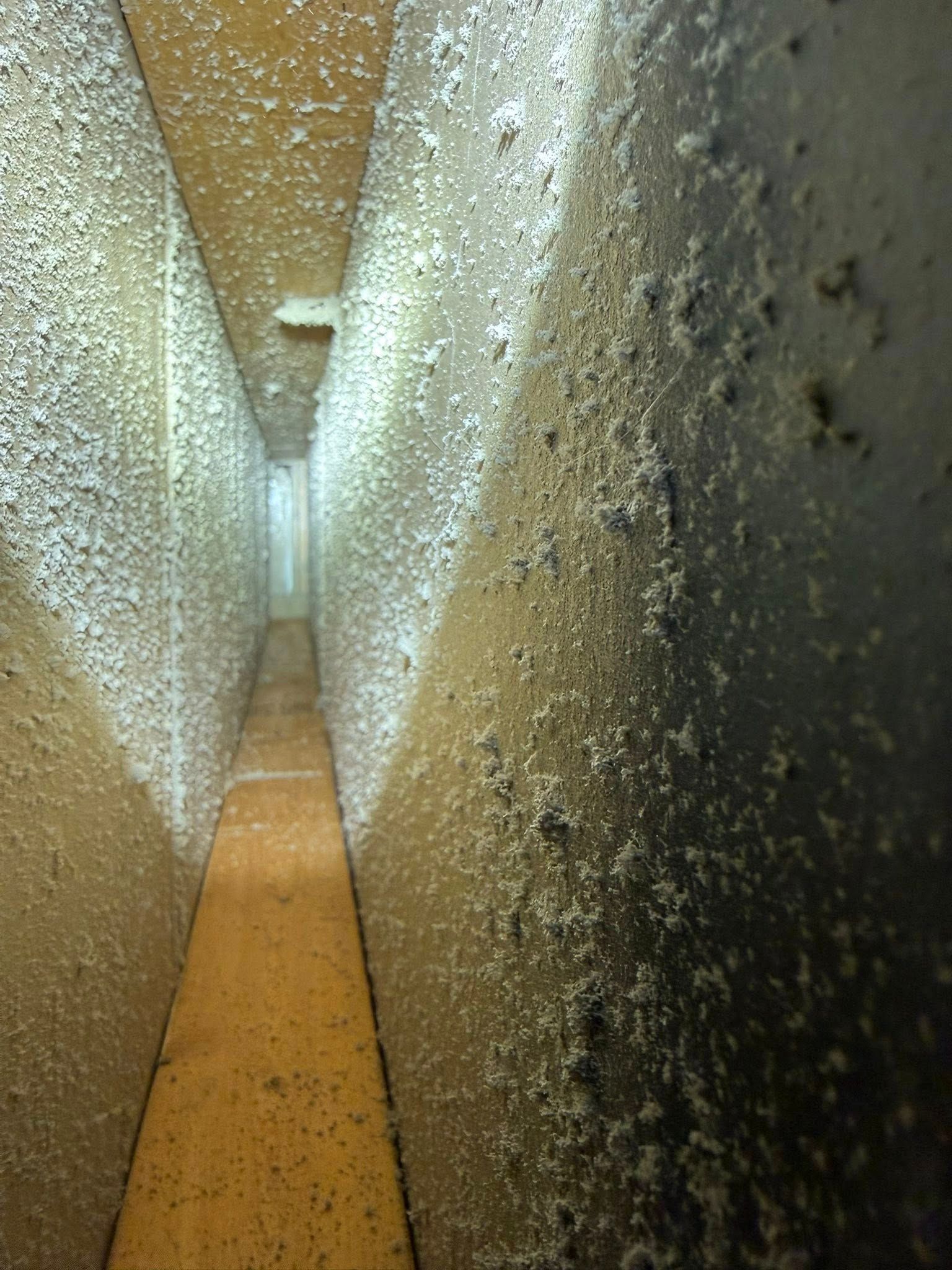 Close-up of a narrow gap between rough concrete walls with a tan floor and speckled white-and-black surfaces
