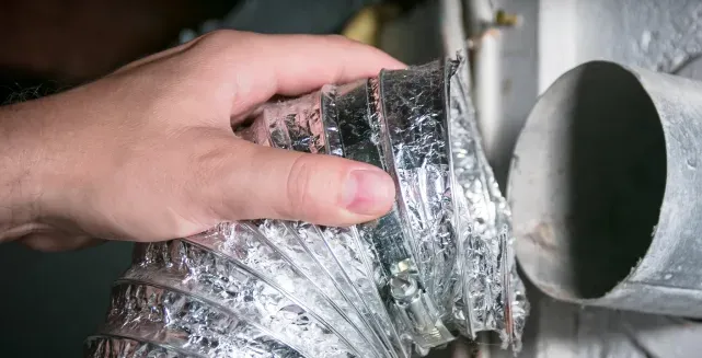 Hand holding a flexible silver HVAC duct near a round metal vent opening