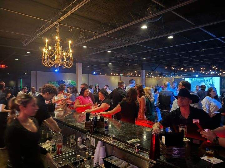 Crowded nightclub bar with people chatting under warm chandelier lights, drinks on the counter.