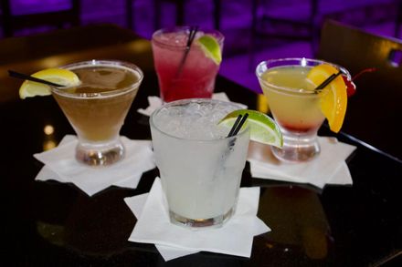 Four colorful cocktails with fruit garnishes on white napkins in a dimly lit bar