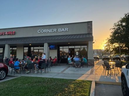 Outdoor corner bar patio with seated diners at sunset, warm lights glowing under a gray building.