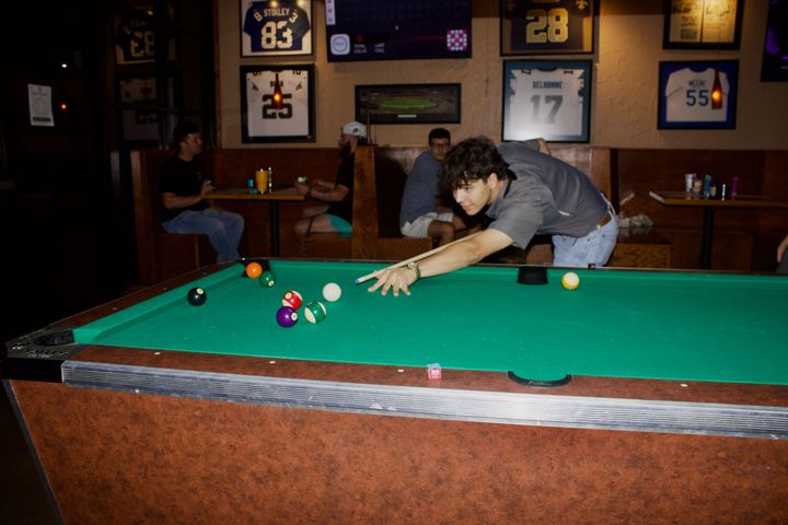 Man leaning over a pool table, aiming a shot in a dimly lit bar with framed jerseys on the wall