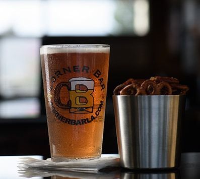 A pint of amber beer beside a metal cup of pretzels on a table in a dim bar.