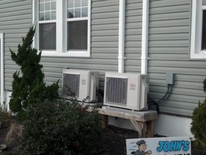 Two Fujitsu AC units on a wooden stand against a gray-sided building with white piping.