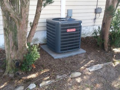 Goodman air conditioning unit on concrete slab between trees near a house.