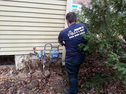 Man in blue shirt trimming branches near a gas meter on the side of a tan house.