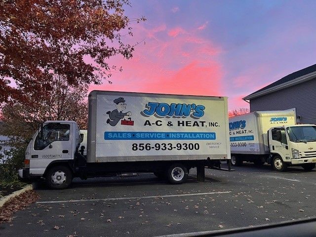 Two white box trucks with “John’s A/C & Heat” parked outside at dusk under pink clouds.