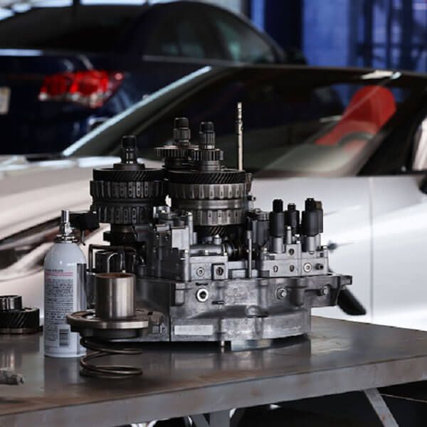 Metal engine component on a workbench in a garage, with a white sports car in the background.