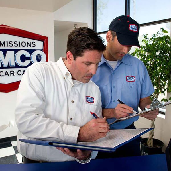 Two mechanics in shirts and caps write on clipboards beside a car service sign