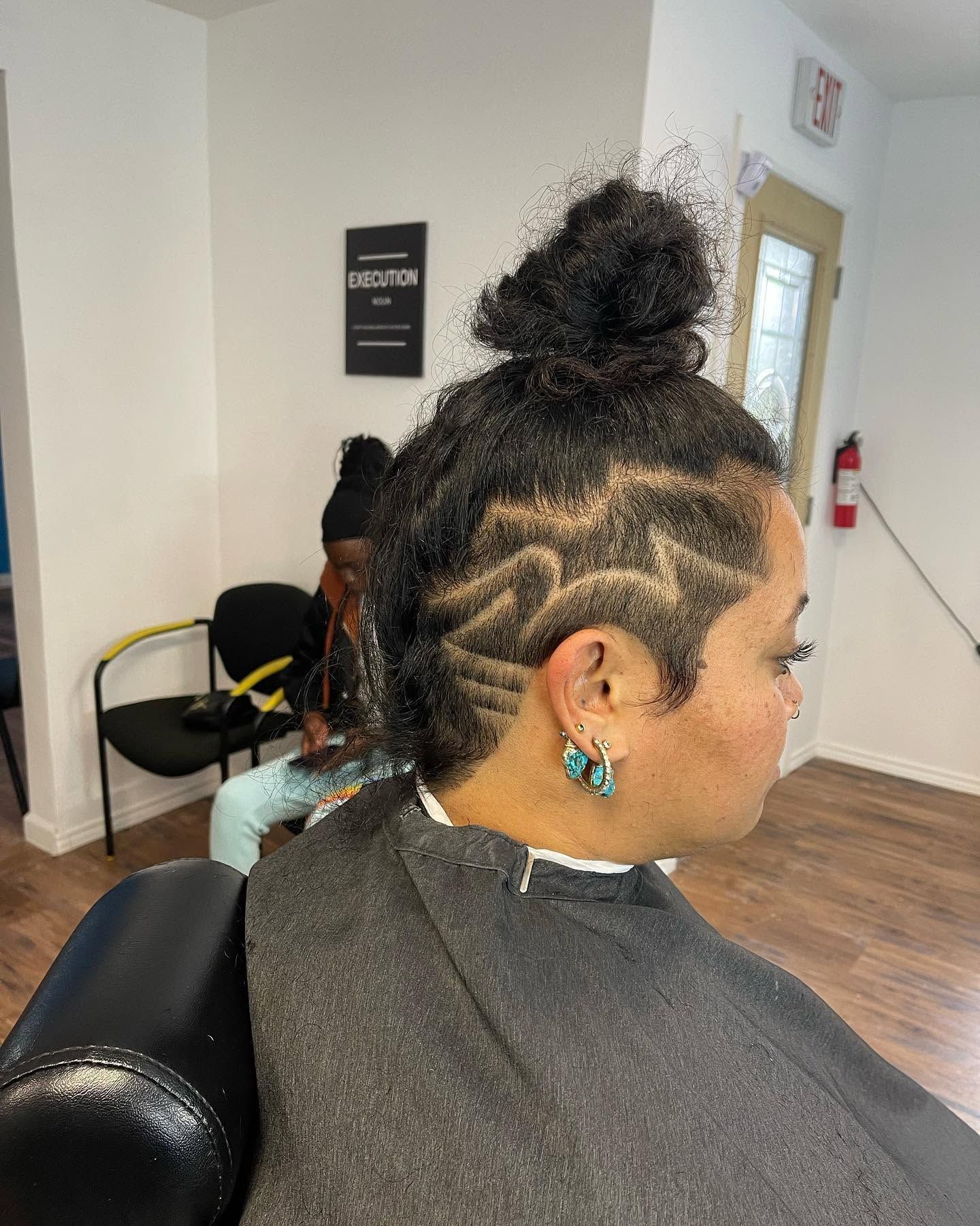 Side view of a braided hairstyle with a high bun in a salon chair, showing shaved zigzag patterns.