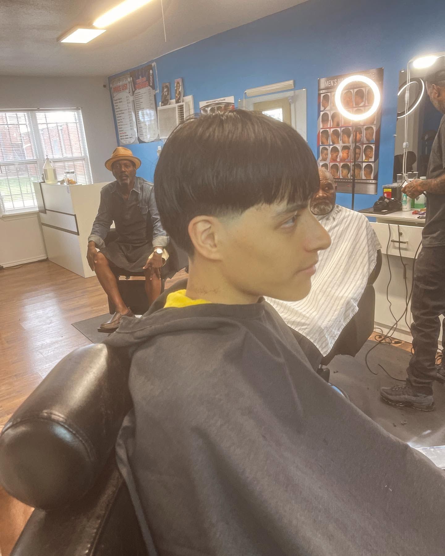 Person with a short haircut sitting in a barber chair, wearing a black cape in a barbershop.