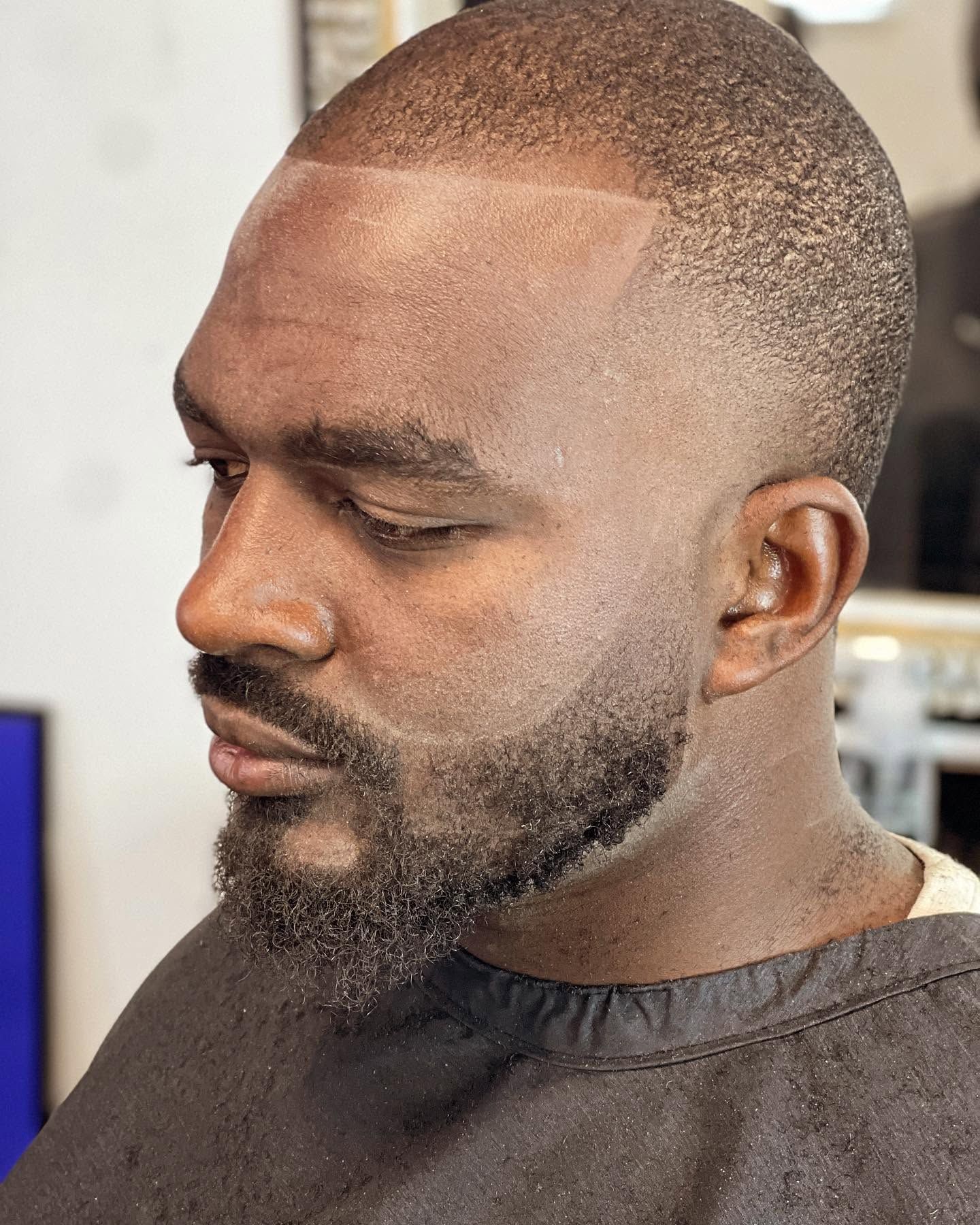 Close-up profile of a man with a short beard and faded haircut, wearing a dark barber cape.
