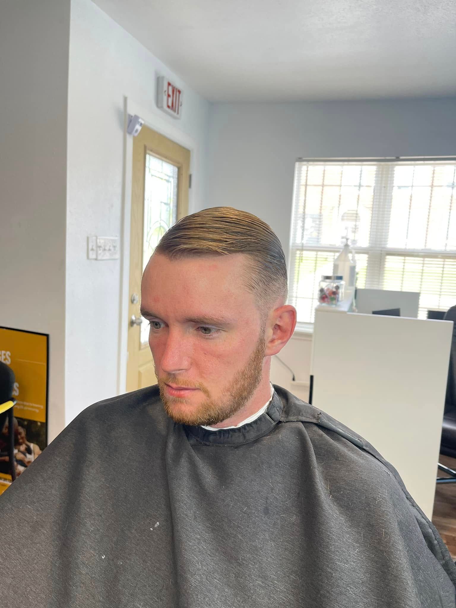Person with short blond hair and beard sits in a barber chair, wearing a cape in a bright salon.