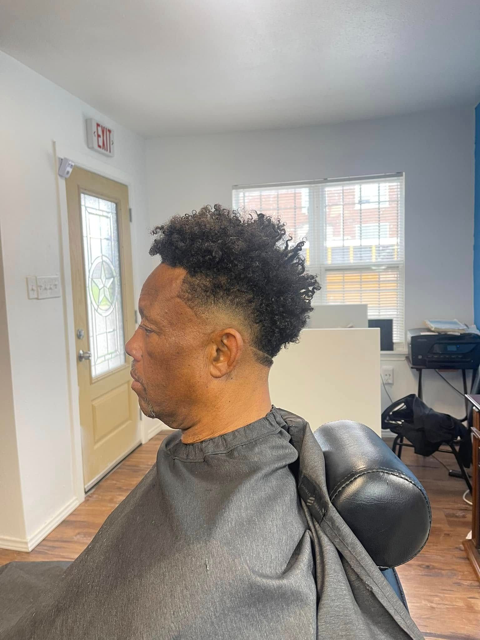 Person in a salon chair, seen in profile with short curly hair, in a bright room with windows and a door.