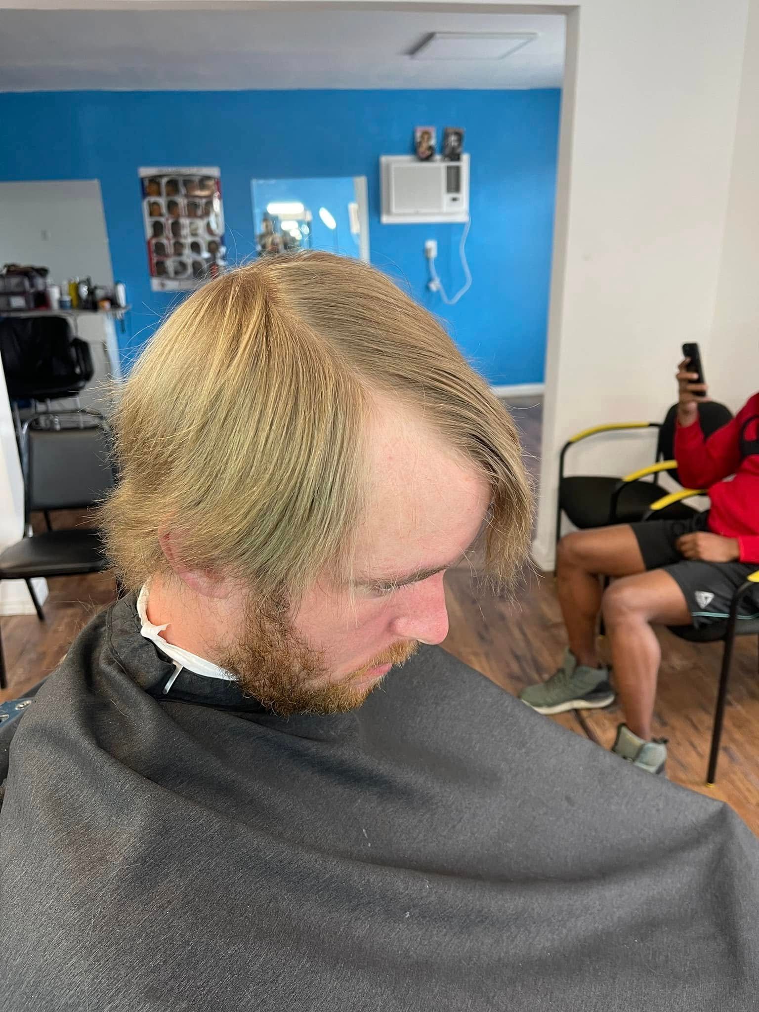 Man in black barber cape with bleached hair sitting in a barbershop, blue wall behind him