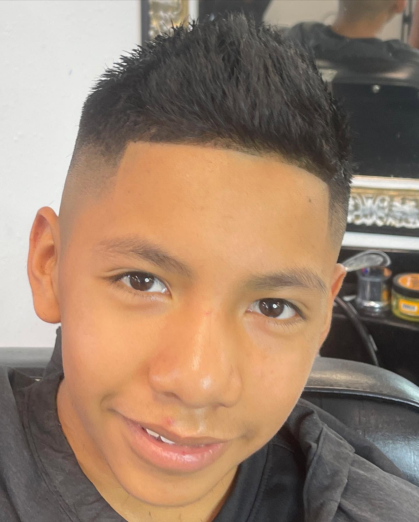 Person smiling with a short black haircut, seated indoors in a barber chair.