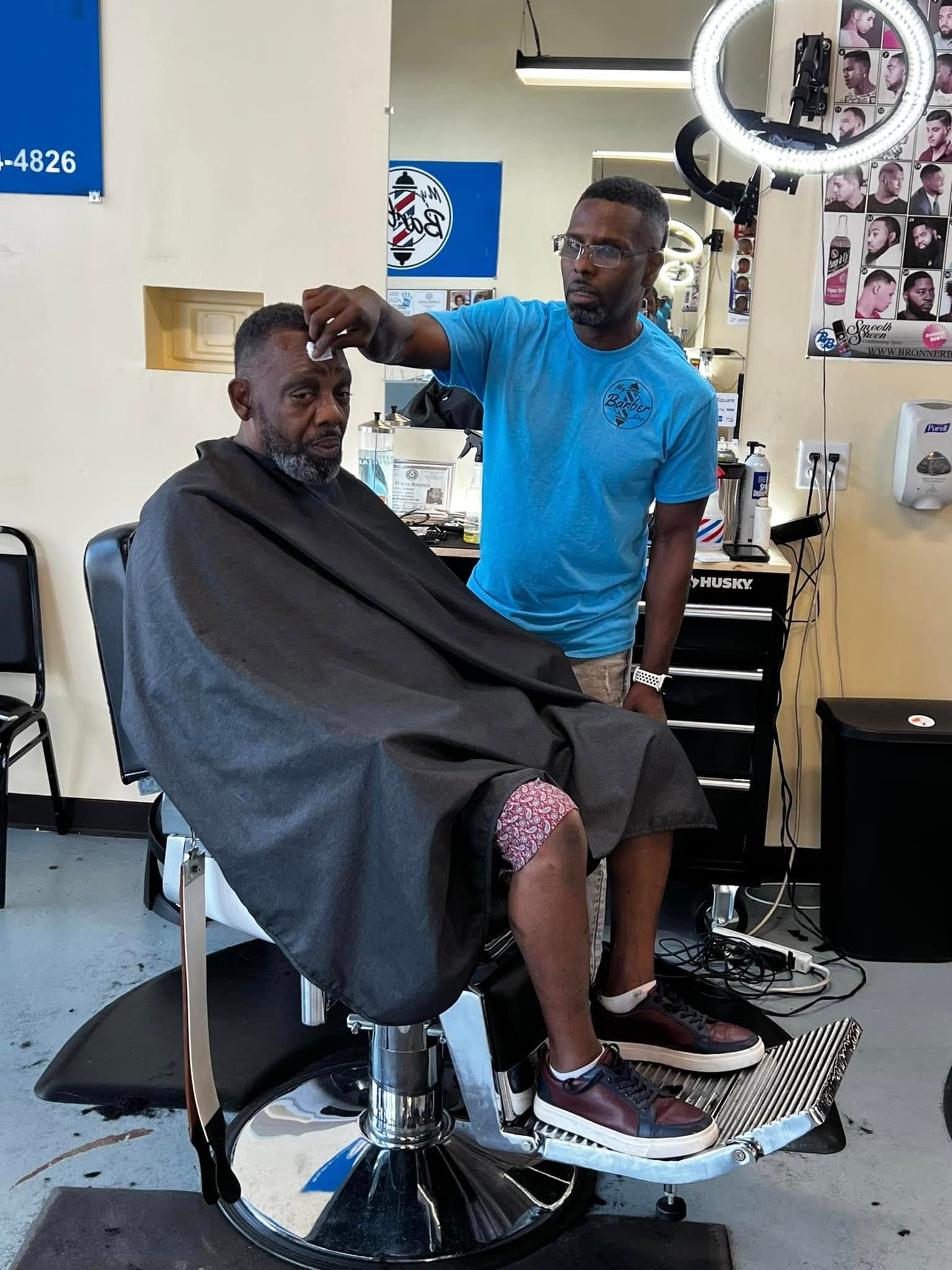 Barber trimming a seated customer’s hair in a barbershop, with a cape and mirror visible.